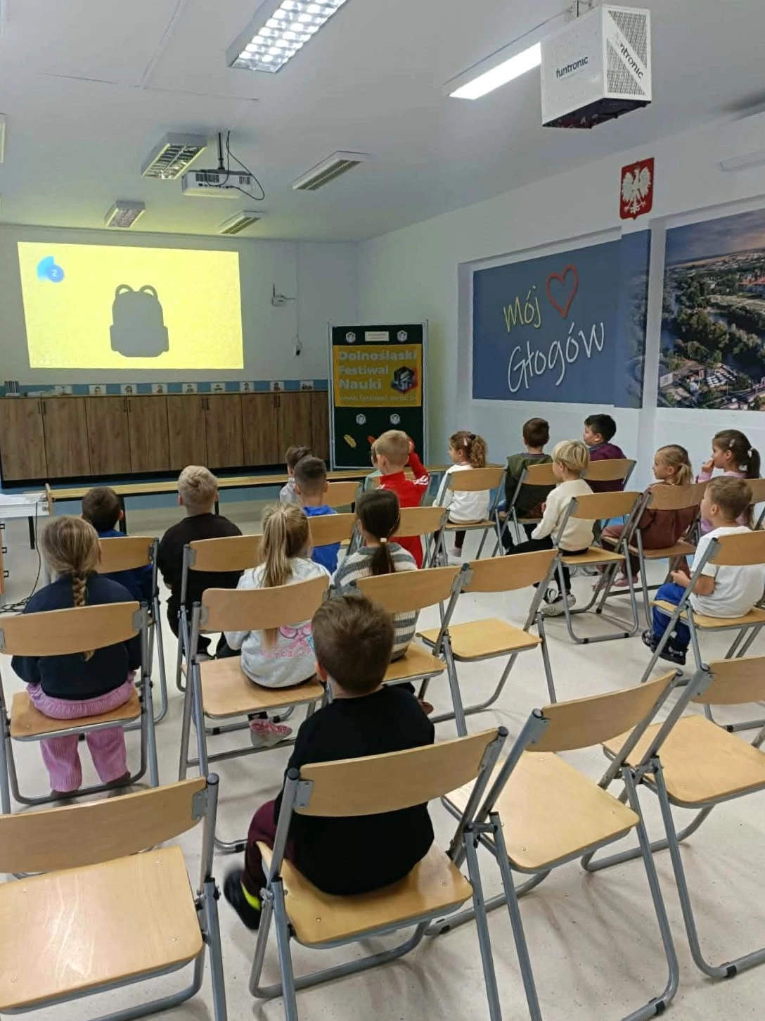 Dzieci siedzą na krzesłach i oglądają prezentację w sali szkolnej.