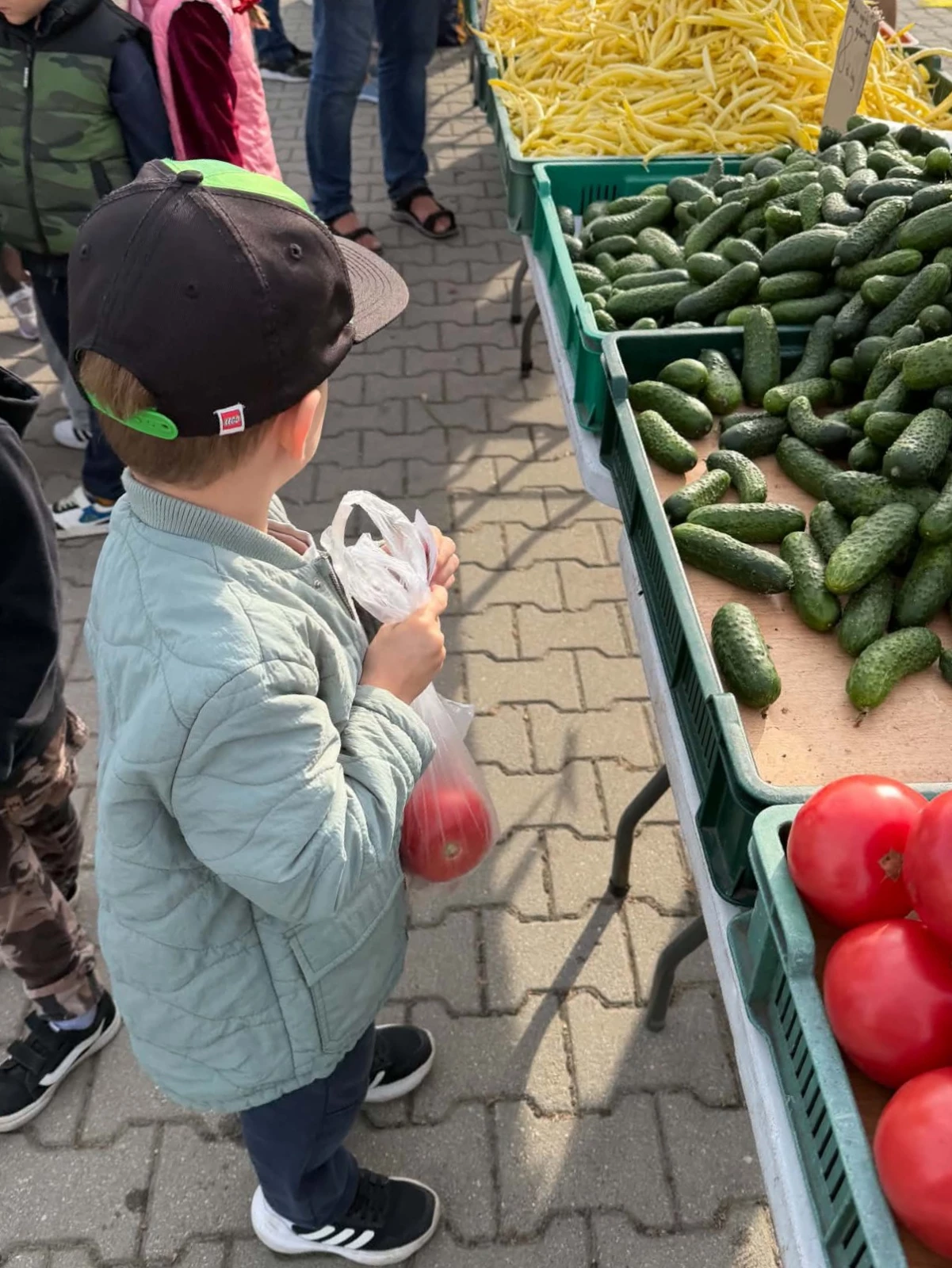 Chłopiec w czapce z daszkiem trzyma siatkę z warzywami przy stoisku.